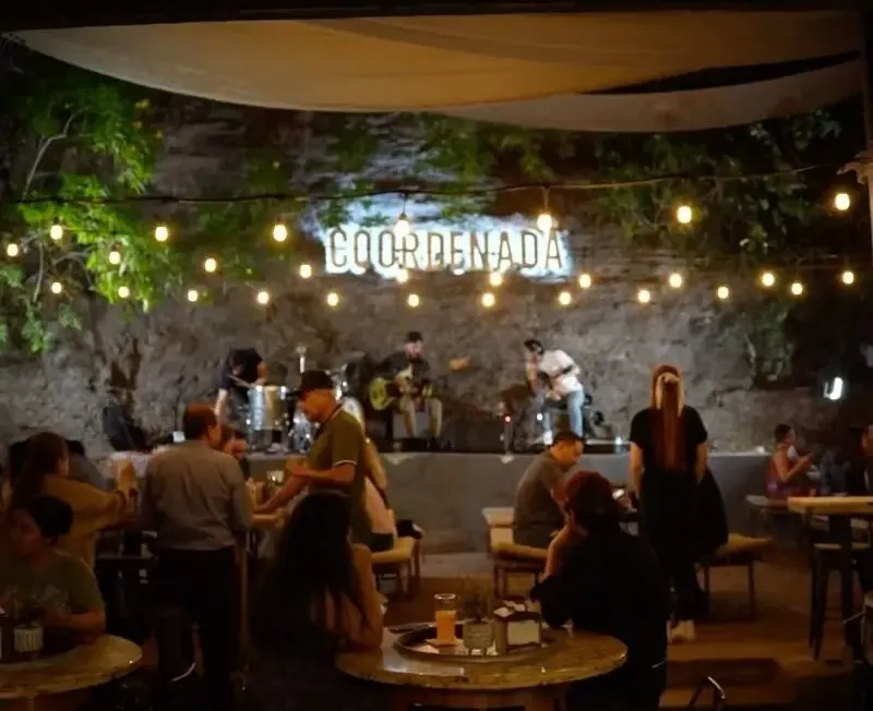 Patio de Cervecería Coordenada de noche, con focos y gente en las mesas